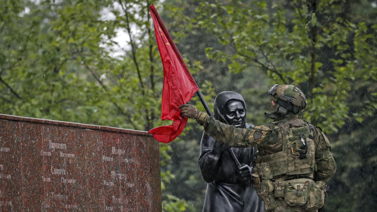 En soldat med ryggen till sätter en röd flagga på en mörkgrå staty av en kvinna.