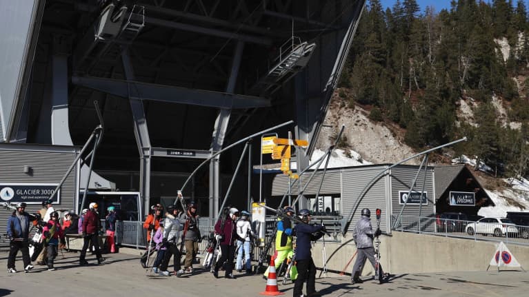 Laskettelijat kävelevät asfalttia pitkin ison hiihtohissin ala-asemalle auringonpaisteessa. Taustalla näkyy paljasta vuorenrinnettä.