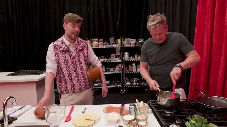 Ruskeahiuksinen nuori mies katsoo, kun vaaleahiuksinen keski-ikäinen mies kokkaa. Nuorella miehellä on silinteri kainalossaan.