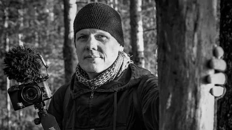 Black and white photo of a man in outdoor clothing and wooly cap holding a camera while leaning on a tree with his hand.