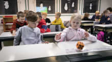 Lapsia istumassa pulpeteissaan koululuokassa. Oikealla lapsi heittää oranssia noppaa. Vasemmalla oleva lapsi tarkkailee vierestä. 
