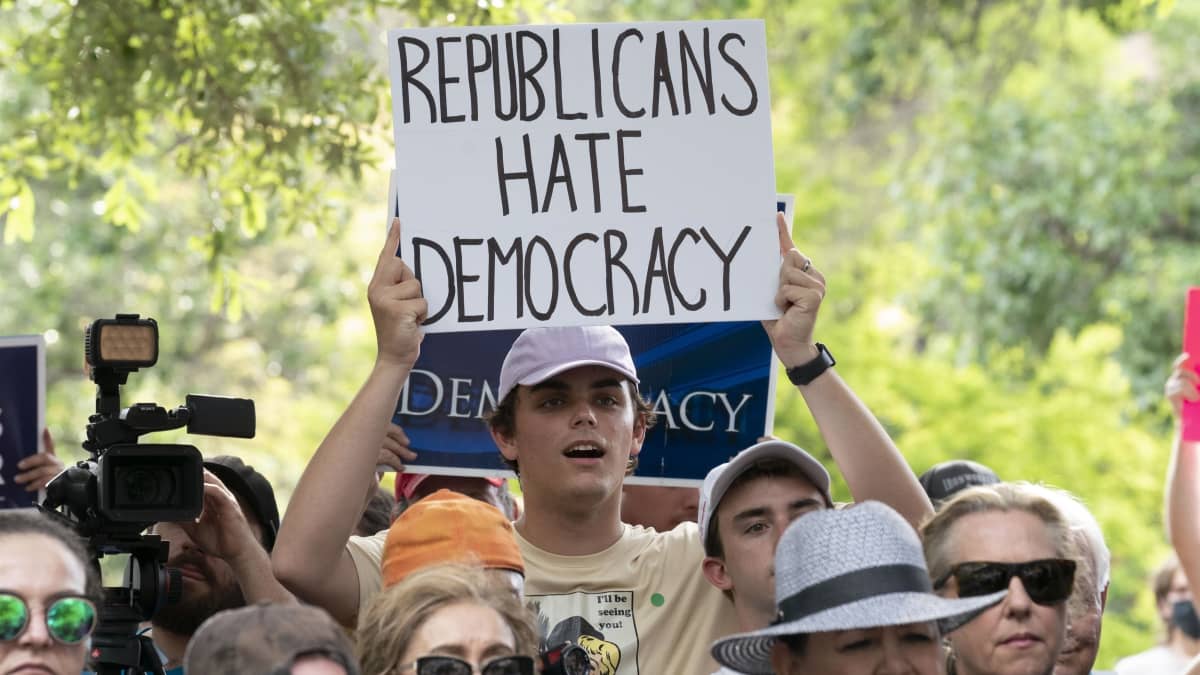 Lippalakkiin pukeutunut valkoinen mies mielenosoittaa. Kyltissä lukee englanniksi "republicans hate democracy".