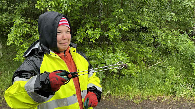 A woman in a rain coat holds up a pair of tongs holding a slug.