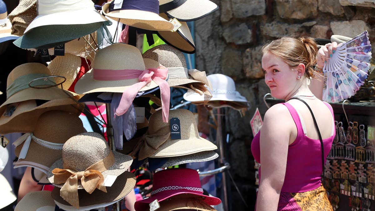 Nainen viuhka kädessään kaupan edustalla esillä olevien hattujen luona.