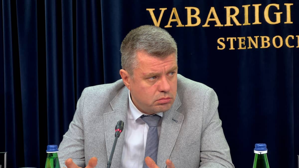 Mies harmaassa pikkutakissa puhuu mikrofoniin pöydän takana istuessaan.