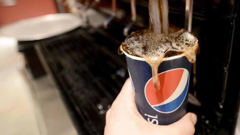 Hand holding a Pepsi-branded blue, red and white paper cup under a soda fountain, being filled with cola.