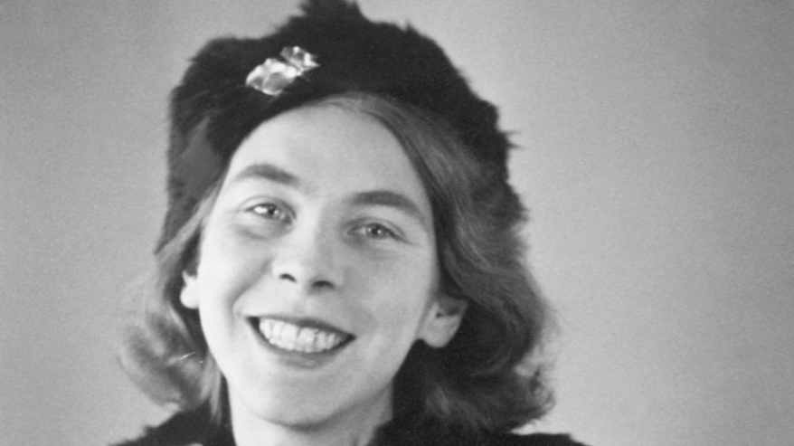 Mustavalkoisessa valokuvassa turkistakkiin ja jalokiveä muistuttavalla koristeella varustettuun turkislakkiin pukeutunut nuori nainen hymyilee leveästi.