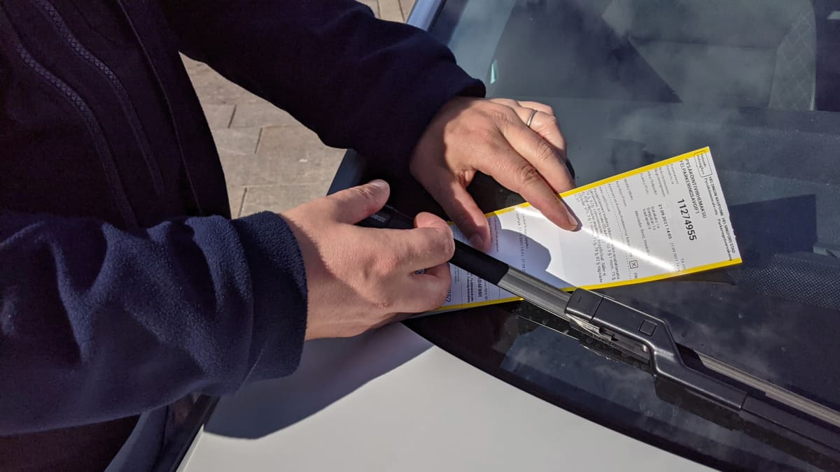 Pysäköinnintarkastaja laittaa pysäköintivirhemaksulappua auton pyyhinsulan alle. Kuvasta on muokattu rekisterinumero pois yksityisyyssuojan vuoksi.