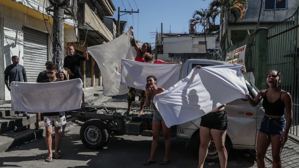 Naiset kannattelevat valkoisia lakanoita favelan kadulla.