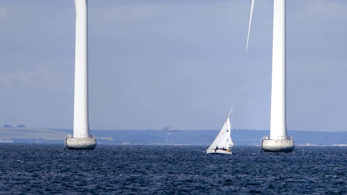 Två stora vindmöllor till havs, med en liten segelbåt mellan sig. 