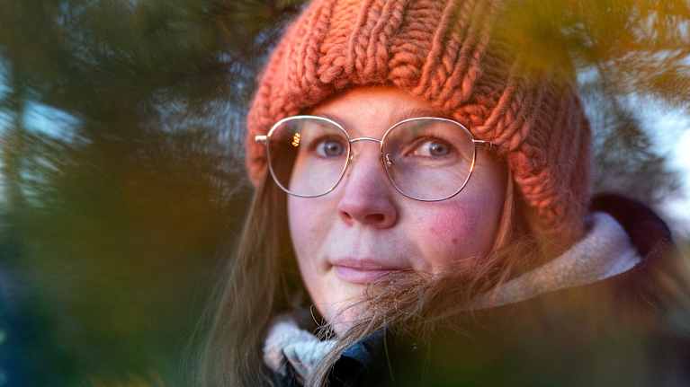 Mari Laaksonen on pukeutunut lämpimästi talvisäähän. Hänellä on päässään oranssi, paksu neulottu pipo. Taustalla näkyy vihreitä havupuun oksia.