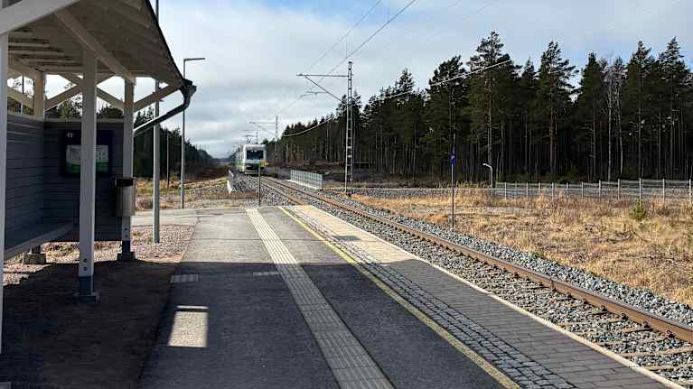 En tågperrong med tak, spår och skog i bakgrunden under en delvis molnig himmel.