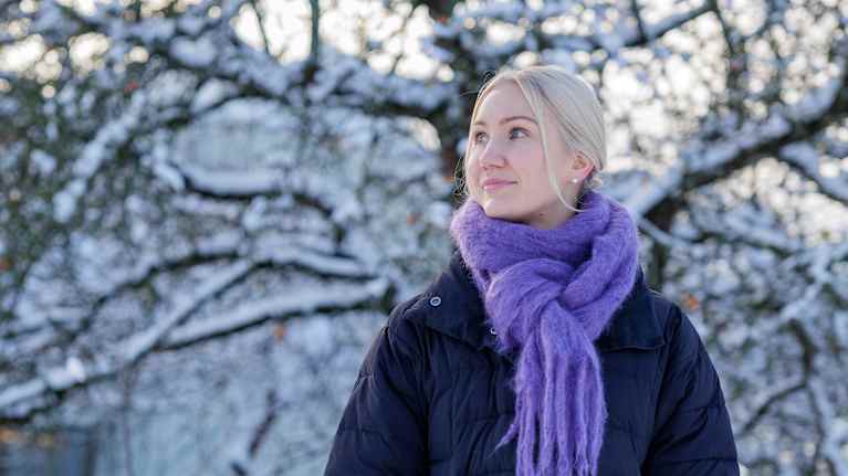 En kvinna iklädd en svart vinterrock och lila halsduk står i en snötäckt park. Hon ler och blickar mot sin höger.
