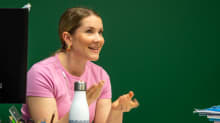 Vaaleanpunapaitainen opettaja Hanna Hallila pöytänsä ääressä liitutaulun edessä.