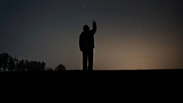 Taivaalle osoittava ihmisen siluetti tähtitaivasta vasten. Korkealla taivallaa näkyy komeetta, kirkas valopallo, jonka perässä himmeämpi pyrstö.