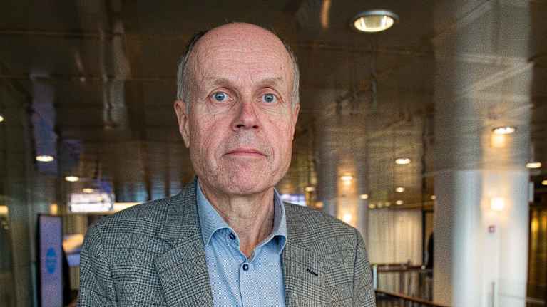 En man i grårutig kavaj och blå skjorta lutar mot ett trappräcke och tittar stint in i kameran.