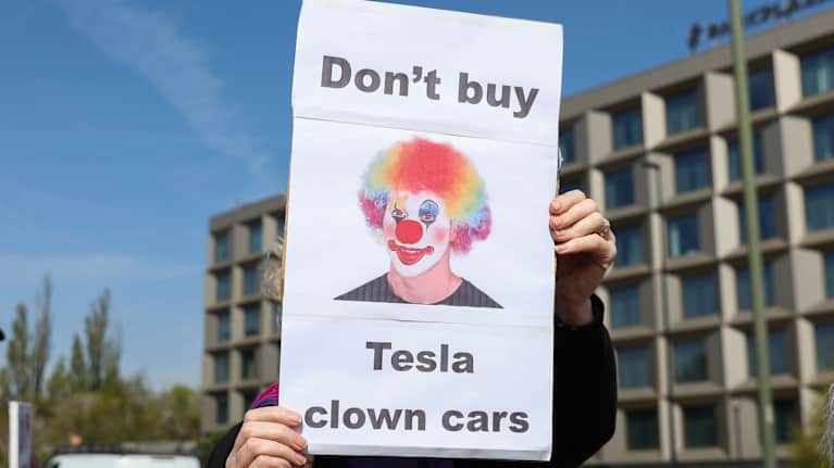 En person håller upp en skylt där det syns en bild på en clown med texten "köp inte Teslas clownbilar".