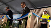 Two men with glasses and dark suits cut a blue-and-white ribbon on a stage while a third man stands smiling behind them wearing yellow hi-vis overalls. 