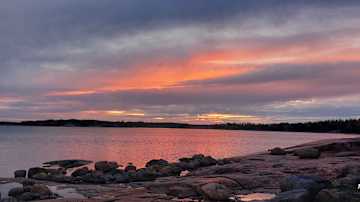 Kaunis auringonlasku merenrannalla, jossa taivasta koristavat oranssin ja violetin sävyt. Kivet meren äärellä luovat rauhallisen tunnelman.
