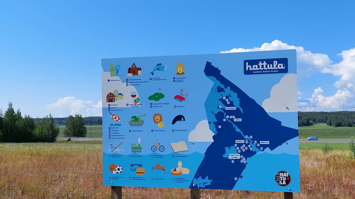 Sinisessä kyltissä on Hattulan kartta. Taustalla viljapeltoa ja taivaalla poutapilviä.