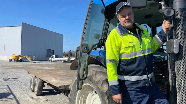 En person står vid en traktor på en byggarbetsplats, iklädd arbetskläder.