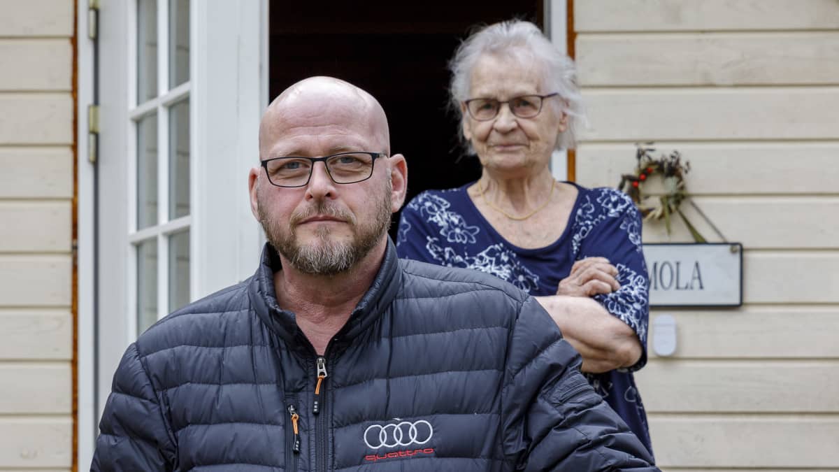 Janne Pulkkinen nojaa kaiteeseen talon ulko-oven edustalla. Äitinsä Leena Pulkkinen kuikuilee hymyillen olan takaa oven raosta.