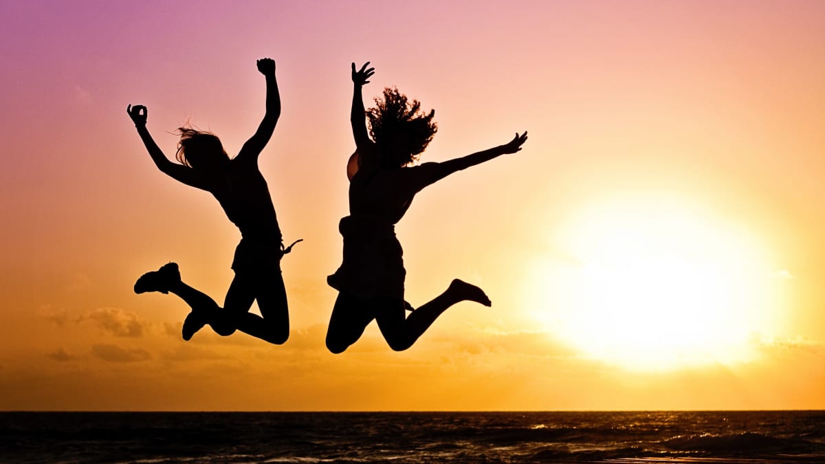 Kaksi ihmistä hyppää ilmaan iloisesti rannalla auringonlaskua vasten