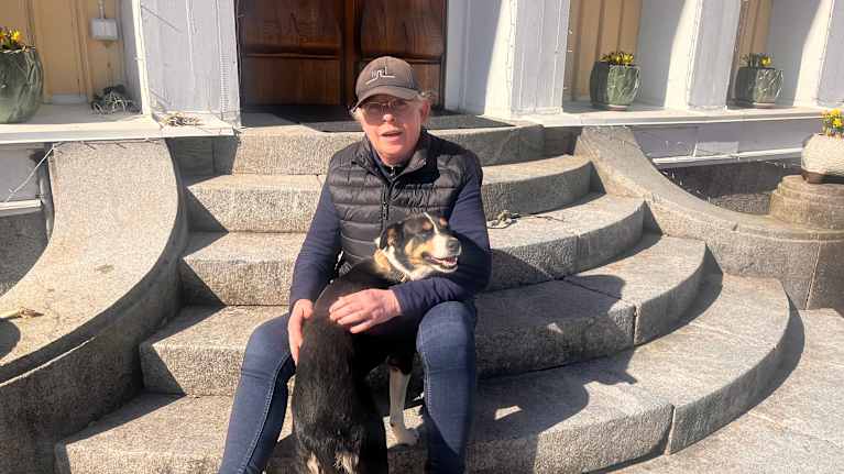 En hund och en kvinna sitter på en stentrappa vid ett gult hus.