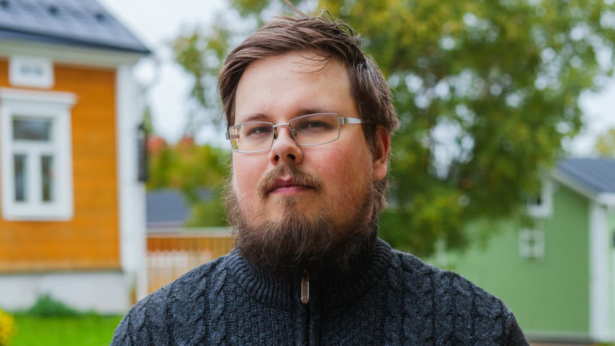 Nuori, parrakas ja silmälasipäinen mies seisoo kadulla ja katsoo kameraan.