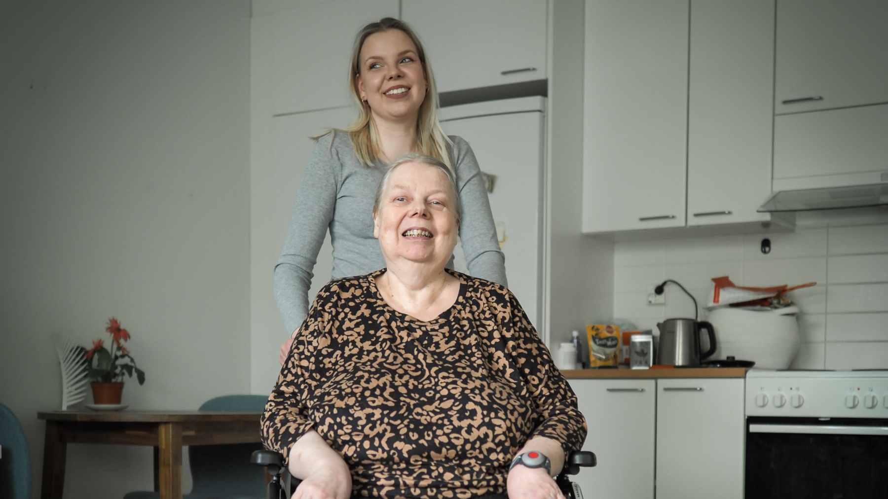 Anne Jurmu istuu pyörätuolissa olohuoneessaan, tytär Iina Hyttinen seisoo hänen takanaan.