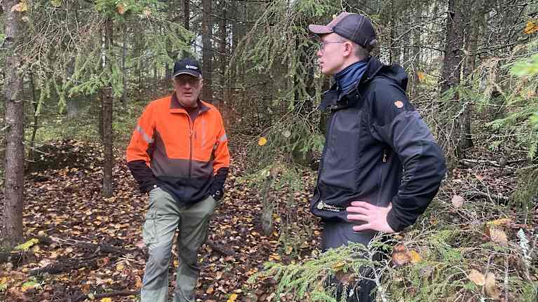 vå personer står i en skog med höstlöv på marken, båda bär kepsar och skogskläder.