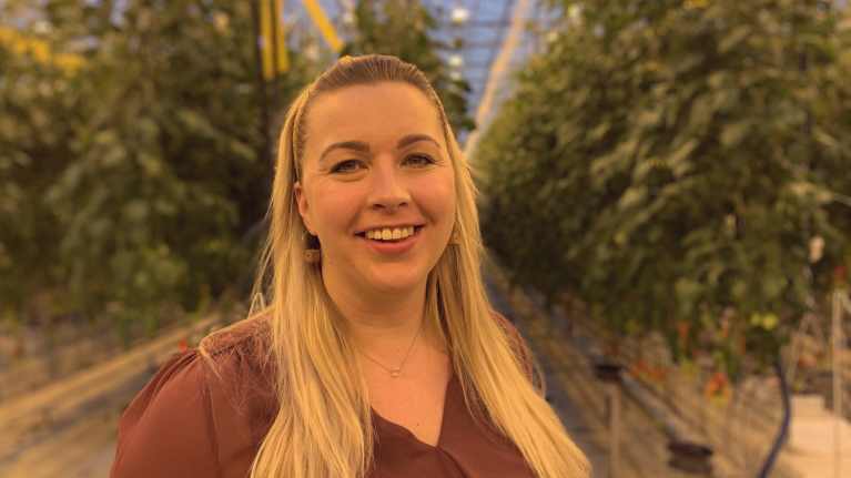 Kvinna med långt ljust hår ler mot kameran. I bakgrunden växthusmiljö med tomatplantor.
