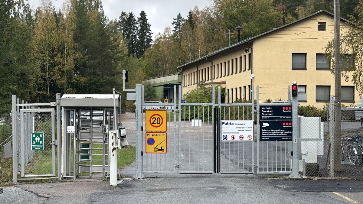 Metalliportin takana on kaksikerroksinen vaalea rakennus, jossa on paljon ikkunoita. Portissa on kyltit, joissa varoitetaan valvonnasta.