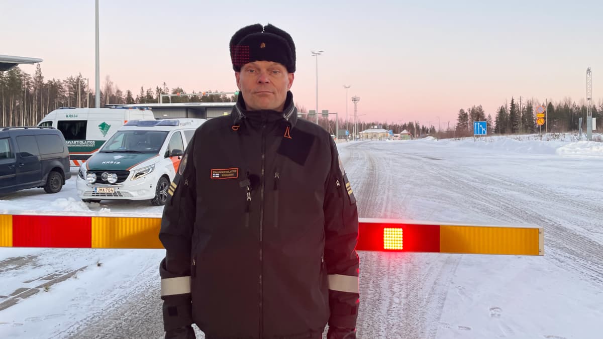 Rajavartiolaitoksen kapteeni Jouko Kinnunen seisoo rajapuomin edessä ja katsoo kameraan, taustalla talvinen maisema ja partioautoja.