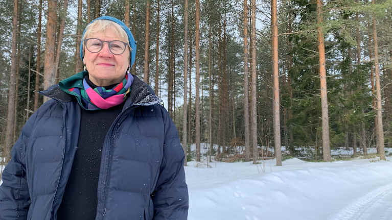 En kvinna med glasögong, blå mössa och uppknäppt jacka står i en vintrig skog. 