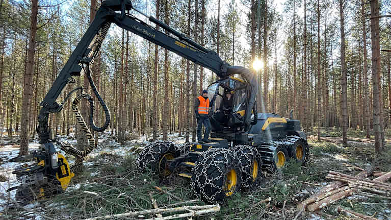 En skogsmaskin arbetar i en skog med en person i orange väst stående bredvid. Det är vinter och marken är delvis täckt av snö.