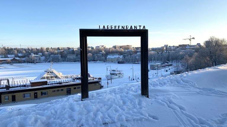 Metallikehikko, jonka päällä lukee Lappeenranta. Taustalla jäinen Saimaa ja luminen satama. 