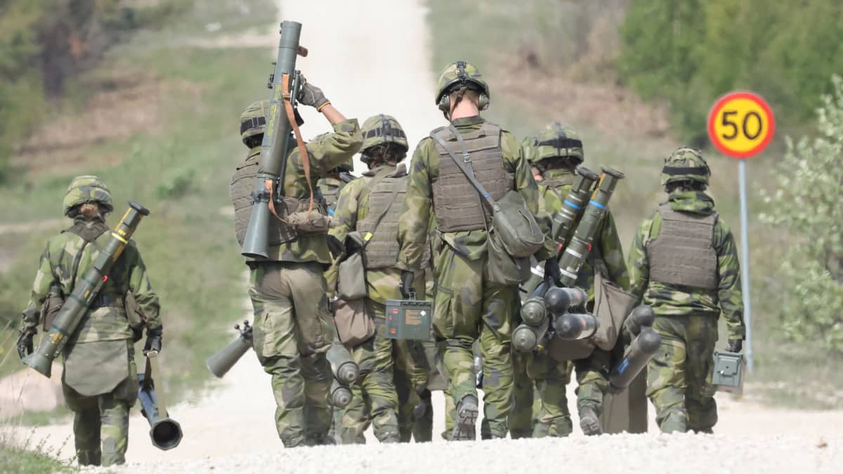 Armeija-asuiset ihmiset kävelevät singot käsissään selin kameraan hiekkatiellä.