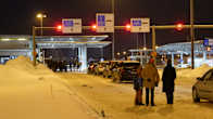 A line of cars and several people, including a child, seen from behind standing in snow at a border checkpoint at night. 