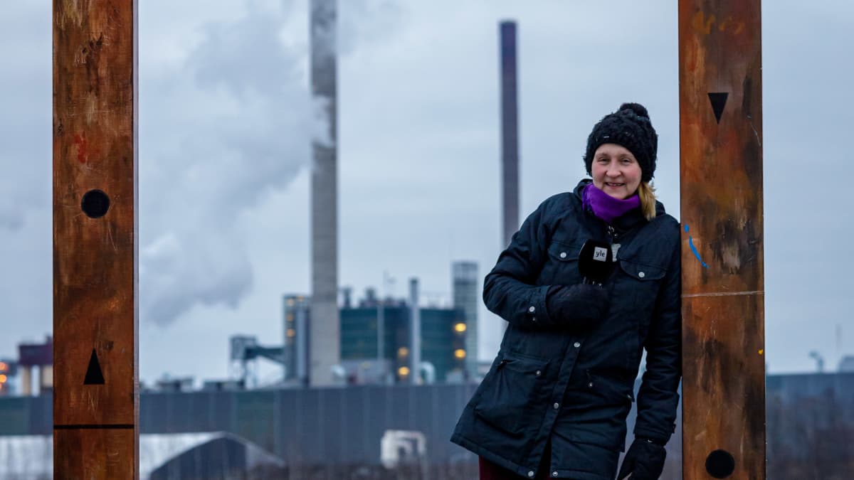 Yle Varkauden toimittaja Tarja Nyyssönen seisoo suuren taulukehyksen sisällä. Taustalla järven takana näkyy Stora Enson tehdas.