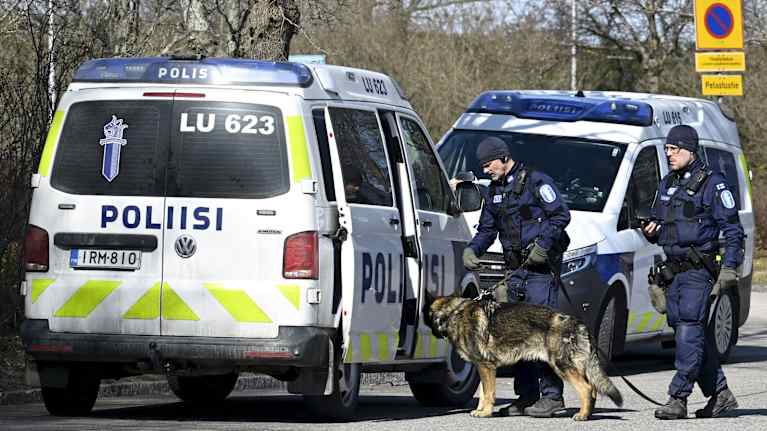 Kaksi poliisia ja poliisikoira poliisiautojen vieressä.