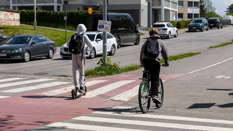 Nuori kulkemassa sähköpotkulaudalla ja toinen pyörällä suojatietä pitkin liikenneväylän ylitse.