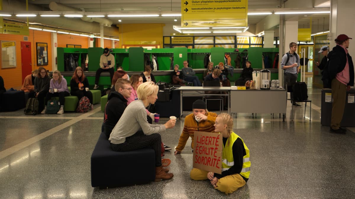 Opiskelijat istuvat Oulun yliopiston aulassa lattialla ja penkillä.