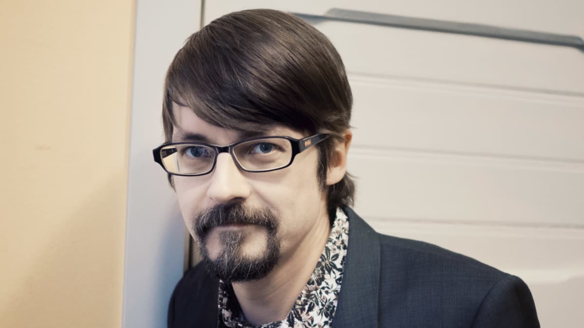 Mies nojaa seinään ja katsoo kameraan.