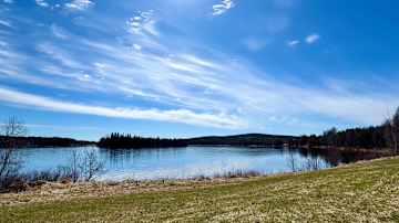 Aurinko paistaa kirkkaalta taivaalta rauhallisen järvimaiseman ylle, kevään merkkejä näkyvissä nurmikolla.