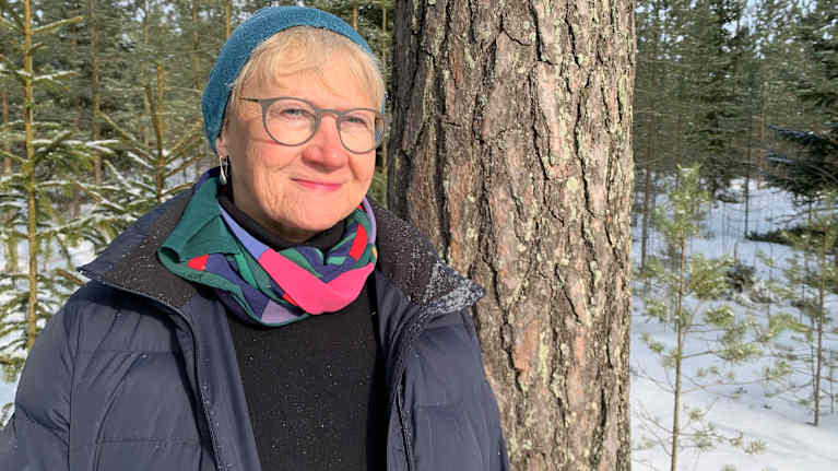 En kvinna med glasögon och blå mössa står med uppknäppt jacka intill en tallstam i en vintrig skog. 