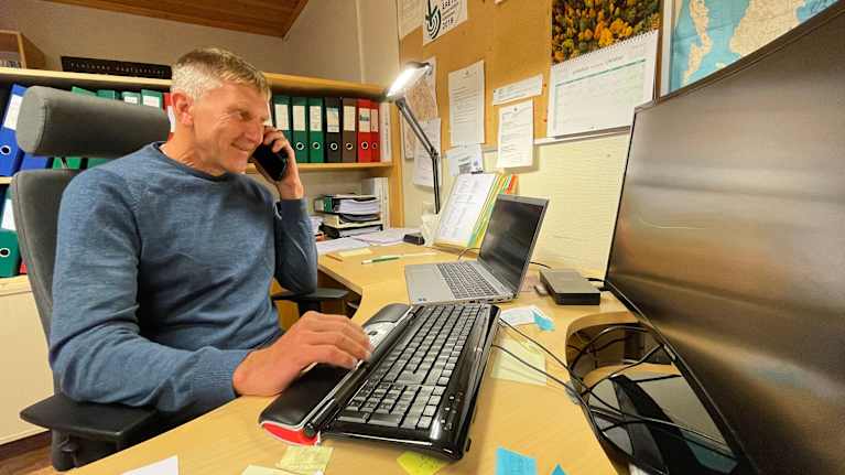 En man sitter vid en dator vid skrivbordet och talar i telefon.