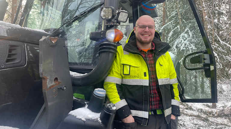 En man med glasögon och skyddskläder står vid dörren till en traktor i en snötäckt skog. 