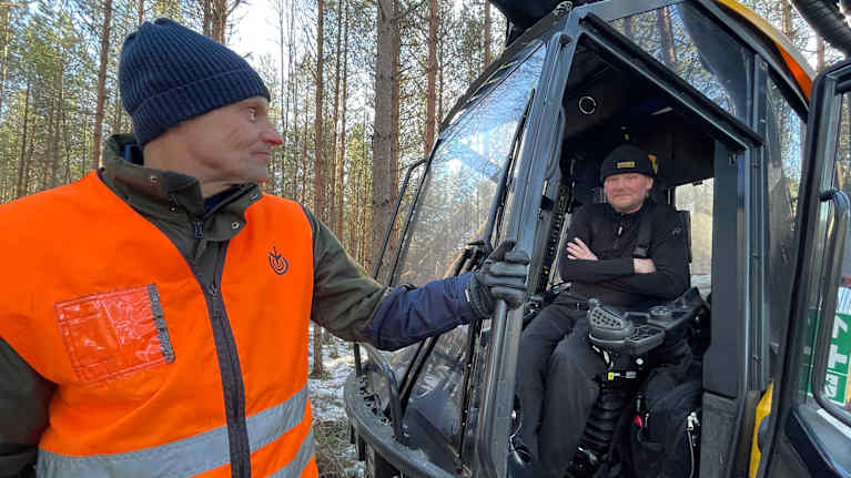 En person i en orange reflexväst står bredvid en skogsmaskin och pratar med föraren som sitter inne i förarhytten.