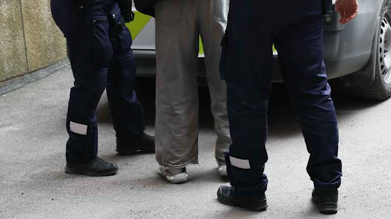 Kaksi poliisia ja nuori mies selin kameraan poliisiauton edessä.